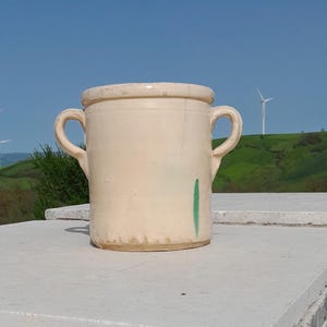 Pode incluir: Um jarro de cerâmica de cor creme com duas alças e uma faixa verde. O jarro está sentado em uma pilha de azulejos brancos.