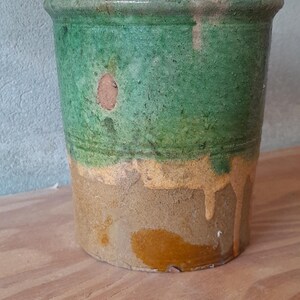 May include: A cylindrical, antique-style ceramic pot with a textured surface. The top half is a vibrant green, transitioning to a band of yellow-orange, and then a tan base. The pot sits on a wooden surface.