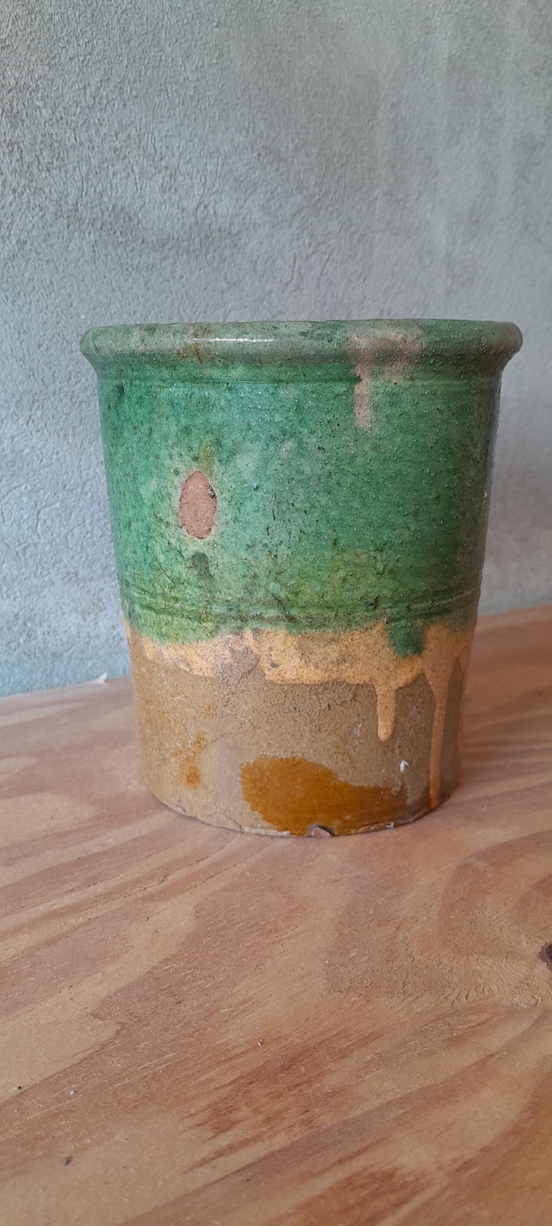 May include: A cylindrical, antique-style ceramic pot with a textured surface. The top half is a vibrant green, transitioning to a band of yellow-orange, and then a tan base. The pot sits on a wooden surface.
