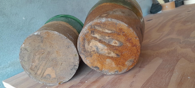 May include: Two cylindrical wooden objects with a weathered, rustic appearance. The smaller one has a green glass top, while the larger one is a darker brown with visible wood grain and a rusty texture. The objects are on a wooden surface.