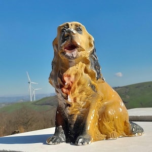 Può includere: Una statua di cane in ceramica gialla e nera con la bocca aperta, seduta su una pila di piastrelle bianche e grigie. La statua è usurata e ha un aspetto rustico. Lo sfondo è un cielo blu con un mulino a vento in lontananza.