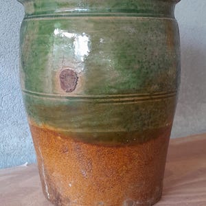 May include: A glazed ceramic pot with a rounded body. The top is green, transitioning to olive green, and then to a warm, orange-brown base. A small, round, brown patch is visible. The pot is likely measured in centimetres.