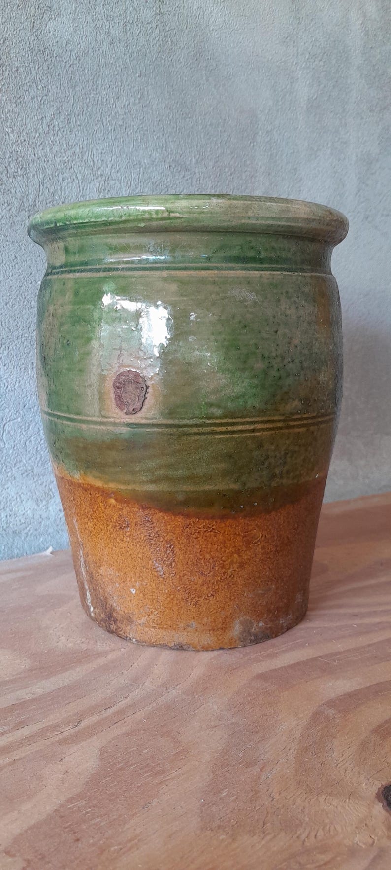 May include: A glazed ceramic pot with a rounded body. The top is green, transitioning to olive green, and then to a warm, orange-brown base. A small, round, brown patch is visible. The pot is likely measured in centimetres.