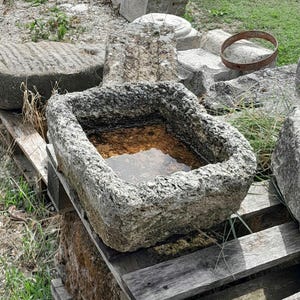 May include: A gray stone trough with a rough texture, filled with water, sitting on a wooden pallet. The trough is surrounded by other stone pieces and green grass.