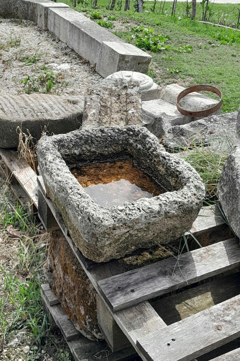 May include: A gray stone trough with a rough texture, filled with water, sitting on a wooden pallet. The trough is surrounded by other stone pieces and green grass.