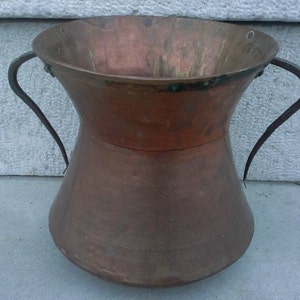 Puede incluir: Un jarr&oacute;n grande de cobre con dos asas de hierro. El jarr&oacute;n tiene una boca ancha y una base estrecha. El cobre tiene un acabado martillado y est&aacute; ligeramente deslustrado.