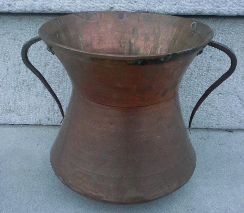 Puede incluir: Un jarr&oacute;n grande de cobre con dos asas de hierro. El jarr&oacute;n tiene una boca ancha y una base estrecha. El cobre tiene un acabado martillado y est&aacute; ligeramente deslustrado.