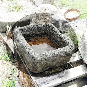 May include: A gray stone trough with a rough texture, filled with water, sits on a wooden pallet. The trough is surrounded by other stone blocks and green grass.