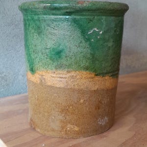 May include: A two-tone ceramic pot. The top half is green, the middle is yellow, and the base is tan. The pot is cylindrical and sits on a wooden surface. The pot is likely for plants.