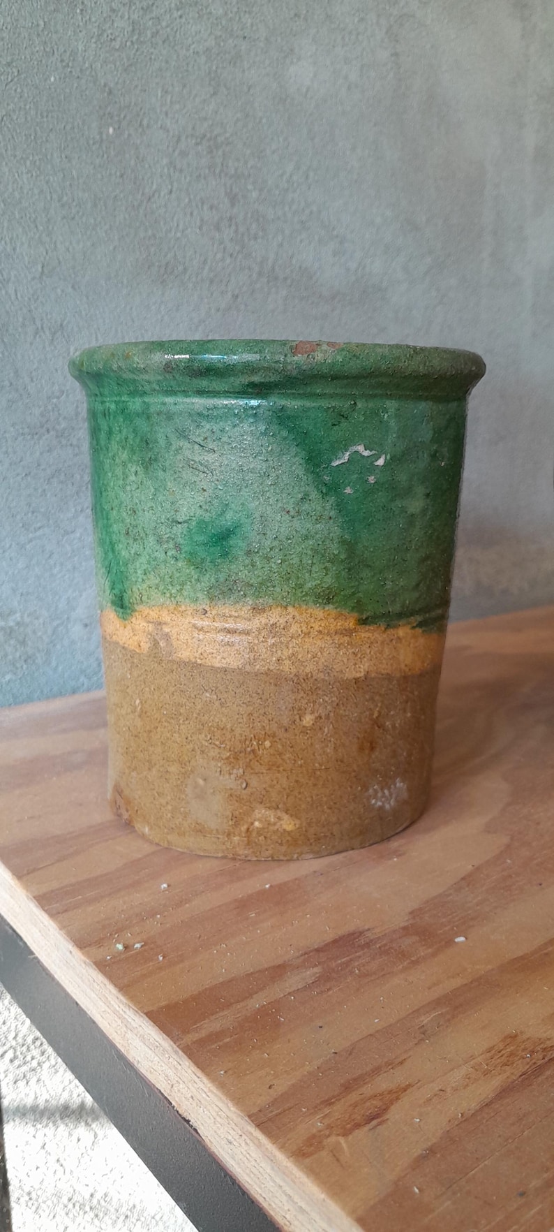 May include: A two-tone ceramic pot. The top half is green, the middle is yellow, and the base is tan. The pot is cylindrical and sits on a wooden surface. The pot is likely for plants.
