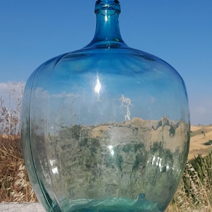 Op de afbeelding: Een grote, heldere glazen fles met een afgeronde body en een smalle hals. De fles heeft een blauwgroene tint en staat op een stenen oppervlak. De achtergrond toont een helderblauwe lucht en droog gras.