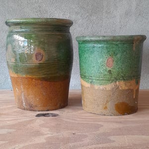 May include: Two rustic ceramic pots. The larger pot is cylindrical with a green top and a brown base. The smaller pot is similar, with a green top and a tan base. Both have a weathered appearance.