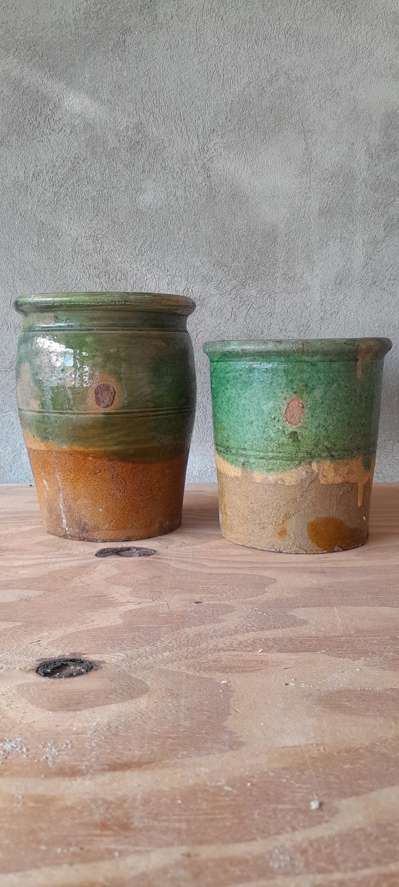 May include: Two rustic ceramic pots. The larger pot is cylindrical with a green top and a brown base. The smaller pot is similar, with a green top and a tan base. Both have a weathered appearance.