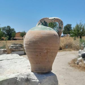 May include: A brown clay jug with a green glaze and a handle. The jug is sitting on a stone surface.