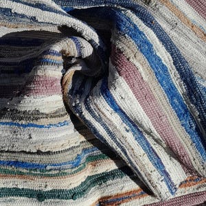 May include: A close-up of a woven rug with a striped pattern in shades of white, blue, pink, and brown. The rug is made of recycled materials and has a distressed, vintage look.