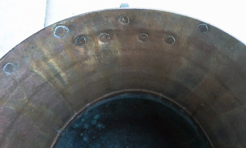 Puede incluir: La parte superior de una olla grande, redonda y de cobre con un interior oscuro. La olla tiene una serie de peque&ntilde;os agujeros redondos alrededor del borde.