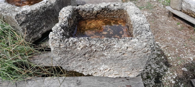 May include: A square, gray stone planter with rough texture and a rectangular opening. The planter is filled with water and has a rustic, weathered appearance.