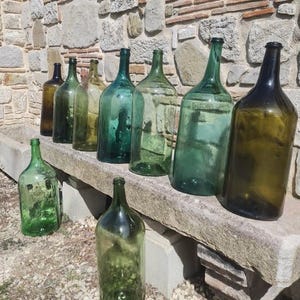 Puede incluir: Una colección de botellas de vidrio vintage en varios tonos de verde y marrón, alineadas en una superficie de piedra. Las botellas son altas y cilíndricas, con un fondo redondeado y un cuello estrecho.