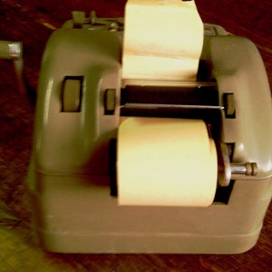 Puede incluir: Una caja registradora vintage de metal gris con un rollo de papel y una palanca en el lateral. La caja registradora tiene un rollo de papel y una palanca en el lateral.