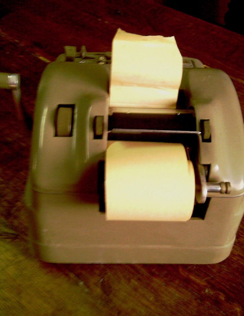 Puede incluir: Una caja registradora vintage de metal gris con un rollo de papel y una palanca en el lateral. La caja registradora tiene un rollo de papel y una palanca en el lateral.