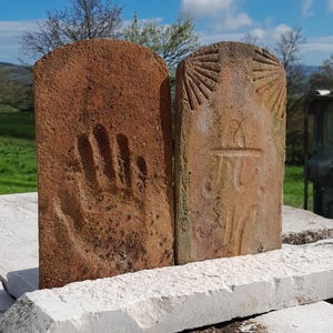 May include: Two terracotta tiles with raised designs. The tile on the left has a handprint, while the tile on the right has a sunburst design and a symbol that looks like a cross.