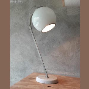 May include: A modern desk lamp with a white globe shade and a brushed metal arm. The lamp is mounted on a circular, light-colored base. The lamp is turned on, illuminating the interior of the shade.