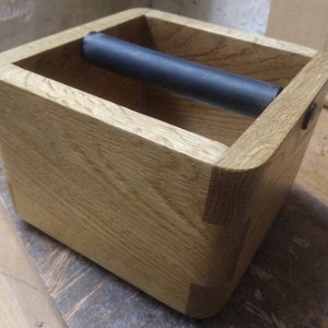 May include: A light brown wooden box with a dark grey cylinder inside. The box has a rectangular shape with rounded corners. The wood grain is visible. The design is simple and functional, suitable for various uses.