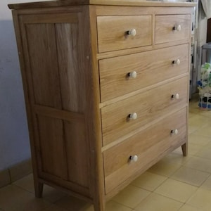May include: A light wood chest of drawers with five drawers and white ceramic knobs. The chest has a single door on the left side.