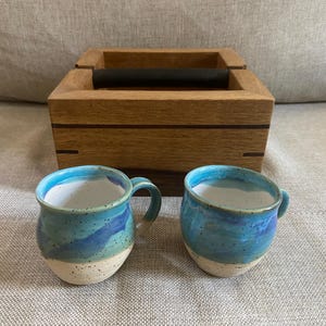 May include: Two blue and white ceramic mugs with handles, sitting on a light brown surface next to a wooden box with a black handle.