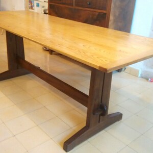 May include: A long, rectangular wooden table with a light wood top and a dark wood base. The table has a simple, sturdy design with a single, wide base.
