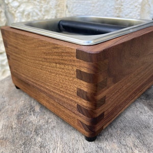 Puede incluir: Un contenedor de café molido de madera con una tapa de acero inoxidable y un asa. El contenedor está hecho de madera oscura con un patrón de veta natural. El contenedor tiene una forma cuadrada y está sentado sobre una superficie de madera.