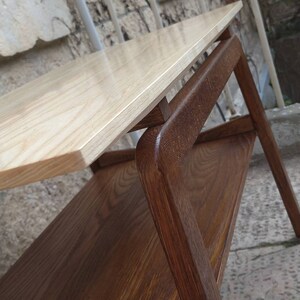 May include: A two-tiered wooden side table with a light wood top and a darker wood base. The table has a simple, modern design with rounded edges.
