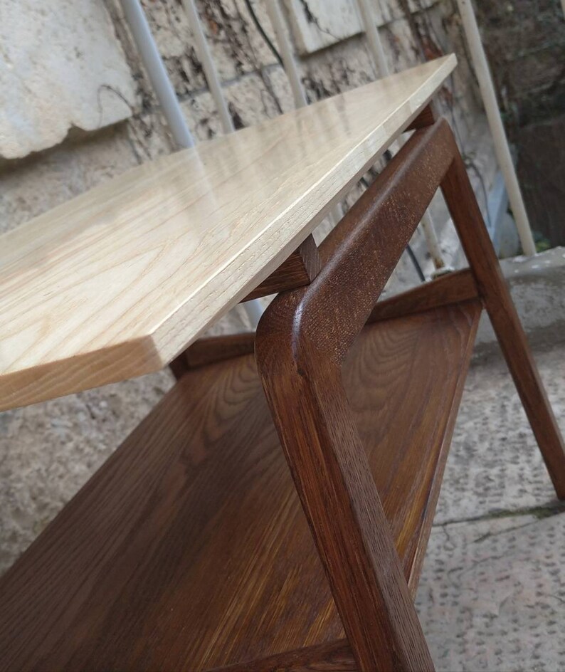 May include: A two-tiered wooden side table with a light wood top and a darker wood base. The table has a simple, modern design with rounded edges.