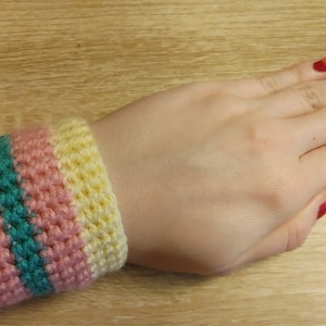 May include: A crocheted wristband in yellow, pink, and teal stripes. The handmade accessory is worn on a wrist. The wristband is made of yarn and has a textured appearance. The nails are painted with a gradient of red, orange, and yellow.