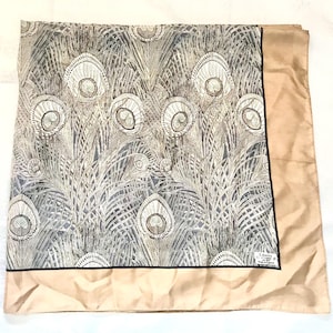 May include: A square scarf featuring a detailed peacock feather pattern in shades of gray, black, and white, bordered by a solid tan edge. A small white label in the corner reads Liberty of London, Est 1875, Made in England, with a black trim.