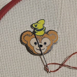 May include: A brown bear with a yellow hat and a needle and thread going through it. The bear is on a beige fabric background.