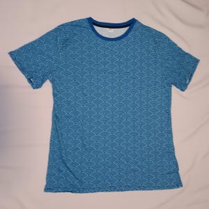 Op de afbeelding: Een blauw T-shirt met korte mouwen met een herhalend patroon van overlappende blauwe en witte halve cirkels.