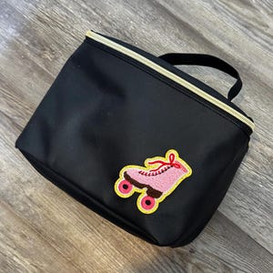 Könnte beinhalten: Schwarze rechteckige Tasche mit goldenem Reißverschluss und kleinem schwarzem Griff. Auf der Vorderseite befindet sich ein rosa Rollschuh-Aufnäher mit gelben und roten Akzenten. Die Tasche liegt auf einer Holzoberfläche.
