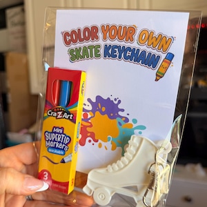 Op de afbeelding: Een transparante verpakking met een witte rolschaatssleutelhanger, een klein doosje Cra-Z-Art Mini Supertip Markers en een kaart met de tekst "COLOR YOUR OWN SKATE KEYCHAIN" en een kleurrijk verfspatterontwerp.