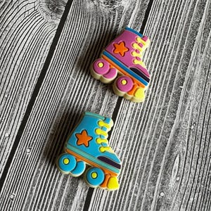 May include: Two colourful roller skate charms. One is pink and the other is blue, both with orange star accents and yellow wheels. The skates have a retro design with a striped pattern and are set against a wood grain background.