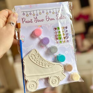 Puede incluir: Un kit de patines de ruedas para pintar en una bolsa de pl&aacute;stico transparente. El kit incluye un pat&iacute;n de ruedas, botes de pintura de varios colores, un pincel y gemas decorativas. El texto "Paint Your Own Create".
