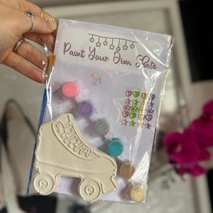 May include: A clear plastic bag contains a paint-your-own roller skate kit. The kit includes a cream-colored roller skate, paint pots in various colors, a paintbrush, and decorative stickers. The text "Paint Your Own Skate" is printed on the packaging.