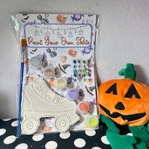 Puede incluir: Un kit de patinaje para pintar en Halloween. El kit incluye un contorno de pat&iacute;n, botes de pintura, un pincel y pegatinas decorativas. Tambi&eacute;n se ve una calabaza de peluche naranja con tallo y brazos verdes.