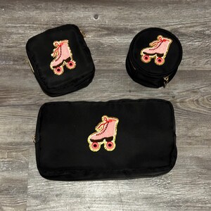 May include: Three black cosmetic bags with gold zips. Each bag features an embroidered pink roller skate design with gold and red accents. The bags are in different shapes and sizes, including a rectangular, a square, and a round bag.
