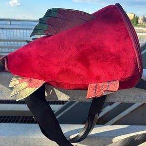 May include: A red velvet fish-shaped backpack with black straps. The backpack features green and yellow fins and a zipper closure. The design is whimsical and unique, perfect for carrying essentials.