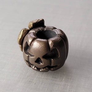 Op de afbeelding: Een donkerbruine keramische pompoenkraal met een gesneden jack-o'-lantern gezicht en een klein gouden blad bovenop.