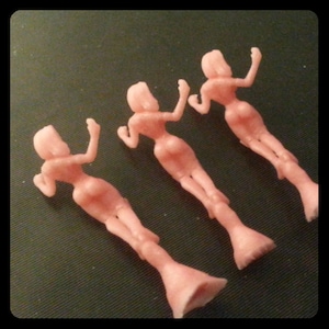 May include: Three pink plastic figurines of women in a standing pose. The figurines are all wearing dresses and have their arms raised above their heads.