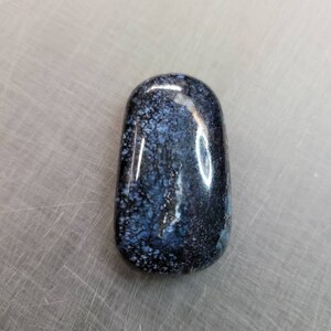 May include: A smooth, polished, dark blue and white stone with a mottled pattern. The stone is oval shaped and has a glossy finish.