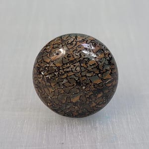 May include: A circular cabinet knob featuring a polished, smooth surface with a complex, dark brown and black mottled pattern resembling fossilized organic material. The knob has a glossy finish that reflects light across its rounded, dome-shaped top.