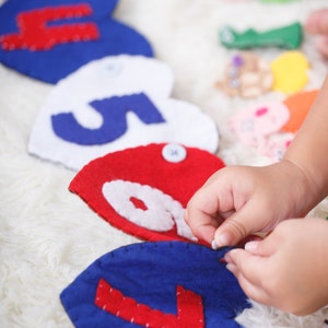 May include: A child's hand is touching a blue felt heart with a red number seven sewn on it. The heart is part of a string of felt hearts with numbers sewn on them. The hearts are in a variety of colors, including red, white, and blue.
